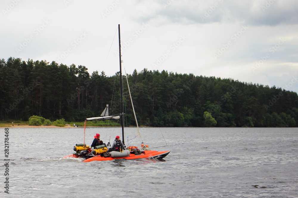Obraz premium An orange inflatable sailing catamaran with a lowered sail floats under the outboard motor on the water against the background of the far green shore on a bad weather cloudy summer day, water tourism