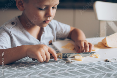 Cute boy 4-5 years old with knife cutting a cheese on the cutting board on the table in the kitchen. Dairy product. Healthy eating and lifestyle.