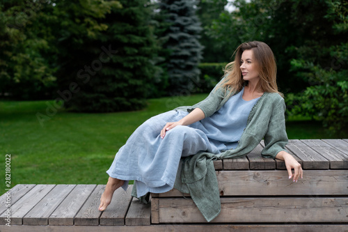 young European girl, natural long hair, beautiful face. blue long dress, gray jacket, sit on wooden platform in green park. Linen womens clothing. hipster lifestyle.