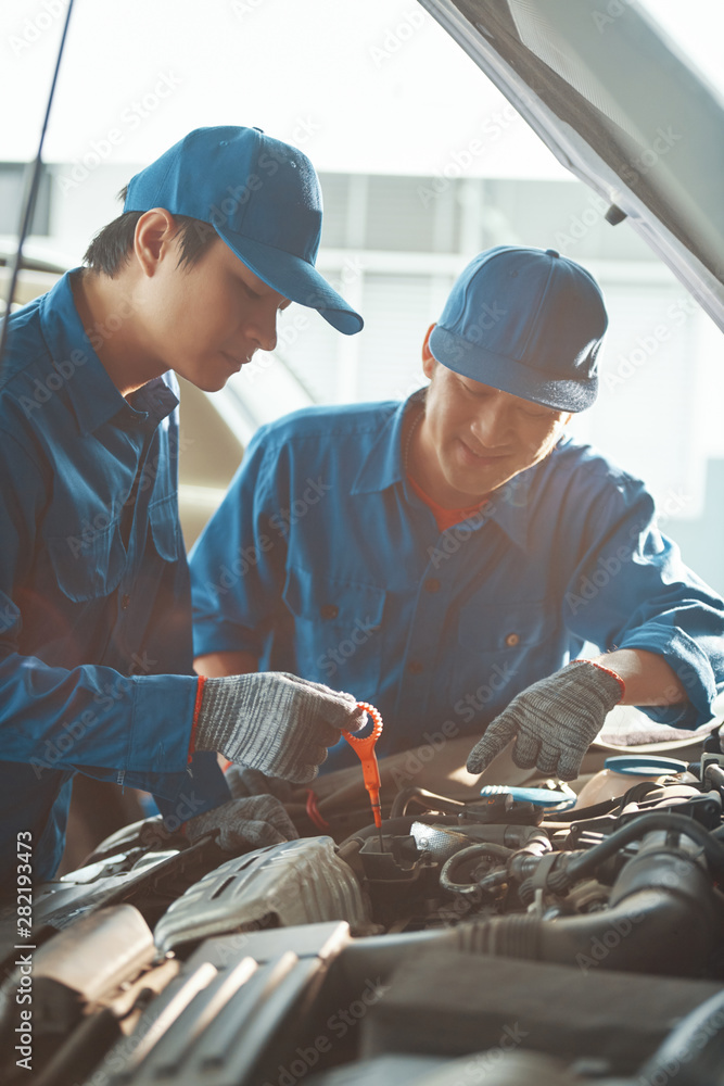 Smiling professional Vietnamese mechanic controlling his coworker checking motor oil level with dipstick Smiling professional Vietnamese mechanic controlling his coworker checking motor oil level with dipstick