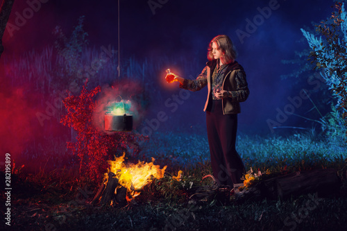 young witch by fire in night forest prepares magic potion