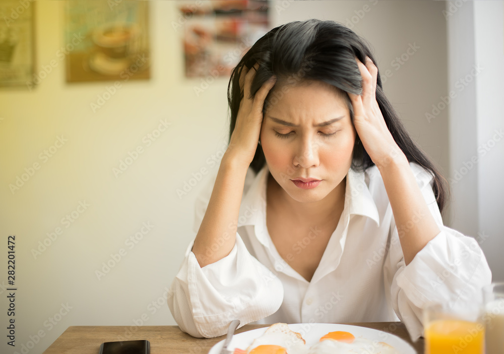 Beautiful Young female in white shirt uses his hands to hold the head to show stress with breakfast on table, Young woman suffering a headache, feeling sick and migraine from tired and health problem. Beautiful Young female in white shirt uses his hands to hold the head to show stress with breakfast on table, Young woman suffering a headache, feeling sick and migraine from tired and health problem.