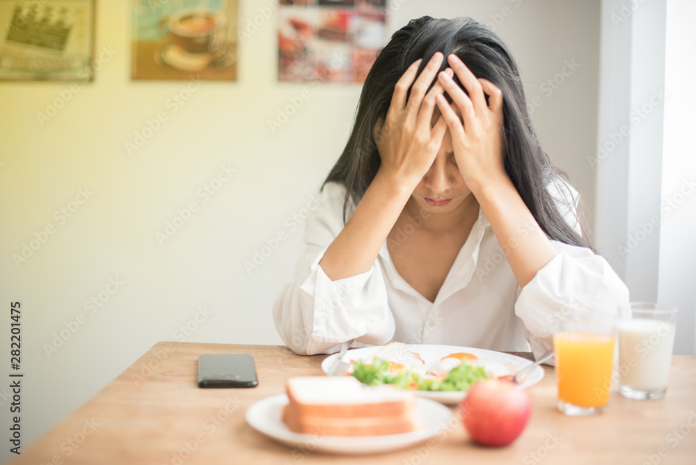 Beautiful Young female in white shirt uses his hands to hold the head to show stress with breakfast on table, Young woman suffering a headache, feeling sick and migraine from tired and health problem. Beautiful Young female in white shirt uses his hands to hold the head to show stress with breakfast on table, Young woman suffering a headache, feeling sick and migraine from tired and health problem.