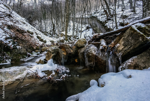 A cold, babbling stream of blue tinted melt water flows after a winter snowstorm. Cascading mountain stream through snow covered woods.Stream in the winter forest. Fairy winter forest with a stream