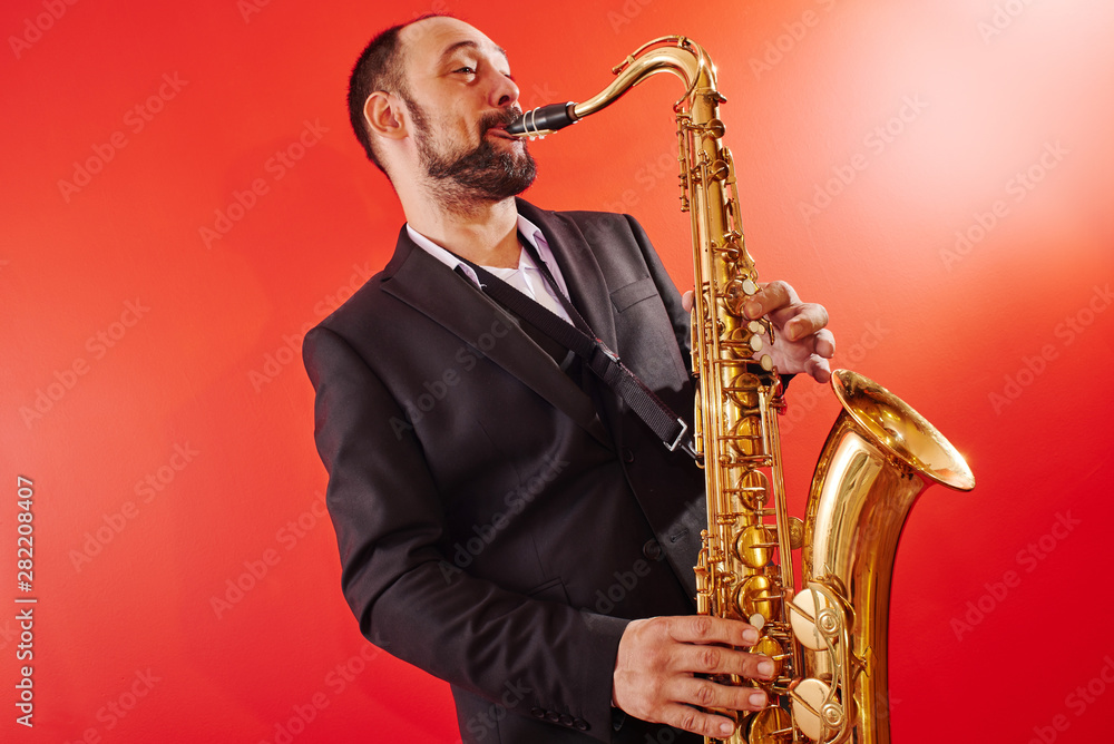 Fototapeta premium Portrait of professional musician saxophonist man in suit plays jazz music on saxophone, red background in a photo studio