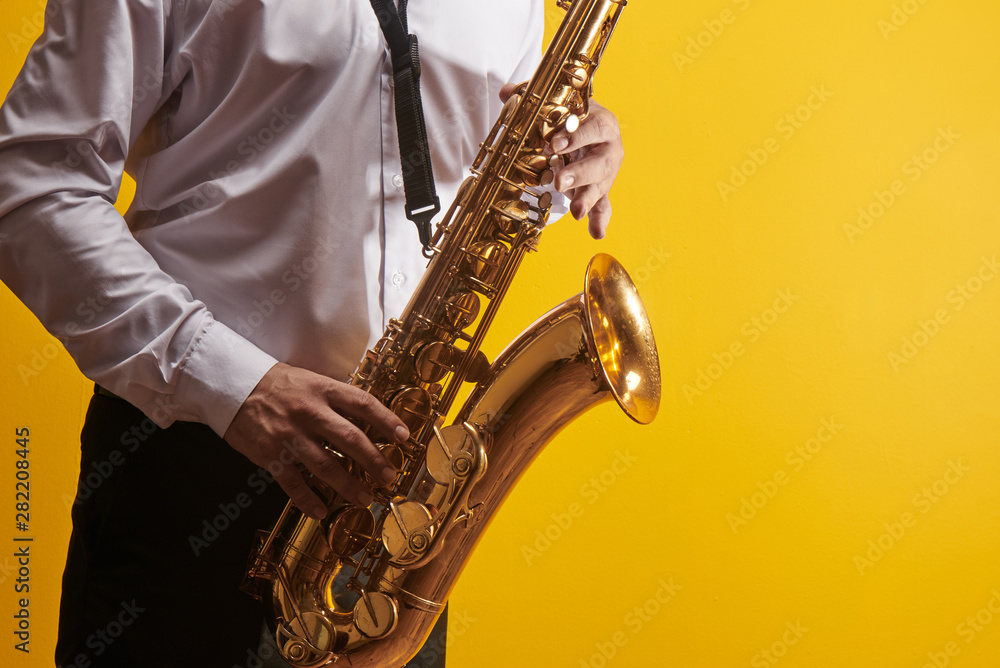 Naklejka premium Portrait of professional musician saxophonist man in white shirt plays jazz music on saxophone, yellow background in a photo studio, side view