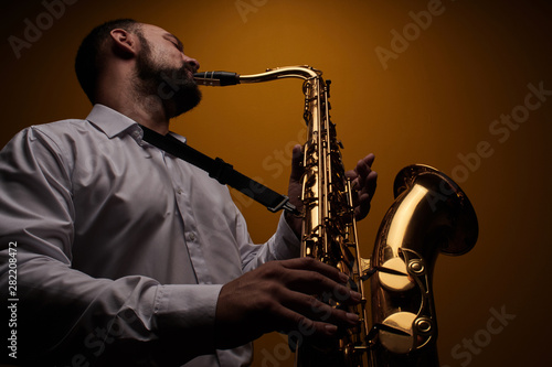 Portrait of professional musician saxophonist man in white shirt plays jazz music on saxophone, yellow background in a photo studio, bottom view