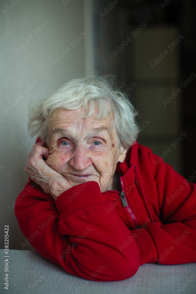 Very old woman in her house, close-up portrait sitting at a table in a red jacket. Plight of pensioners. Very old woman in her house, close-up portrait sitting at a table in a red jacket. Plight of pensioners.