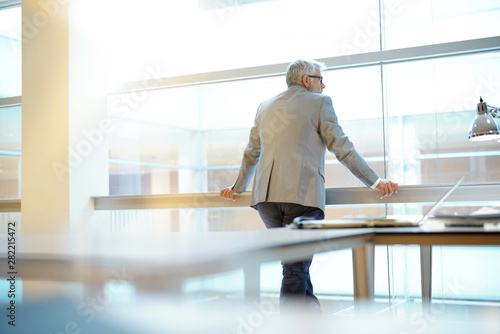 Tableau sur toile Mature businessman looking out large window in office