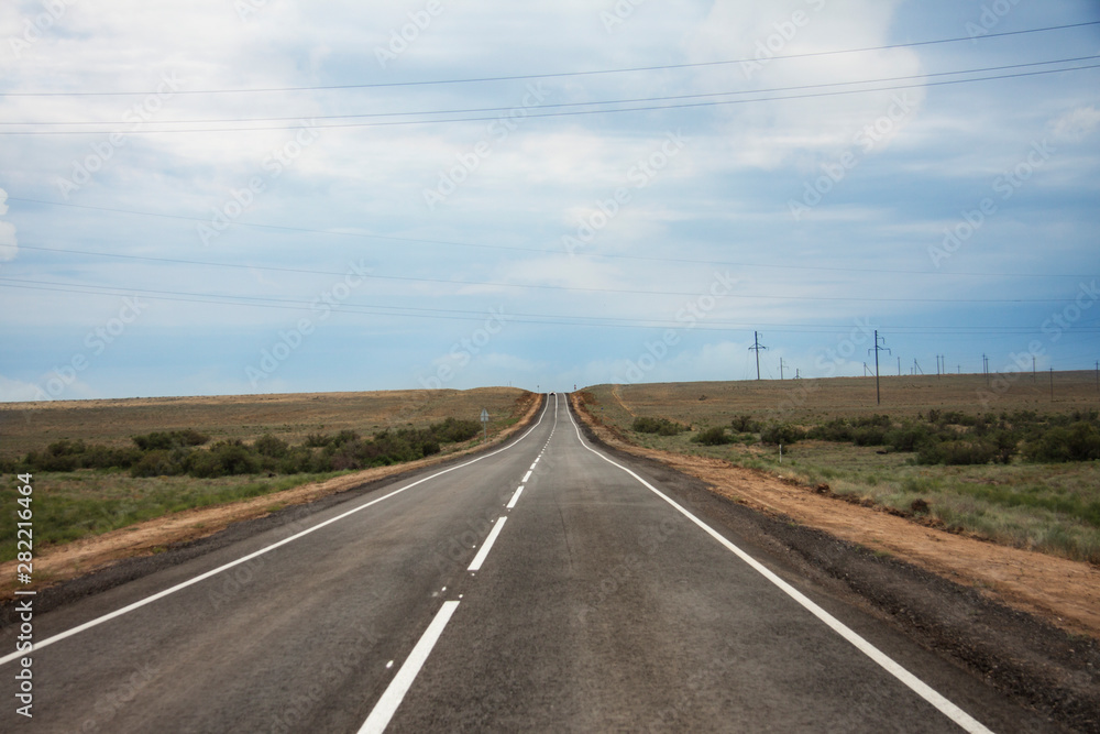 Steppe panorama of a desert landscape summer day. An asphalt road with markings goes forward to the horizon. Traveling among the sands and shrubs. Background image. Dull sky with clouds.