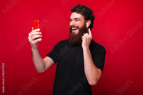 Joyful young bearded man is holding a smartphone in red phonecase and pointing to his eraphones on a red background. © Vulp