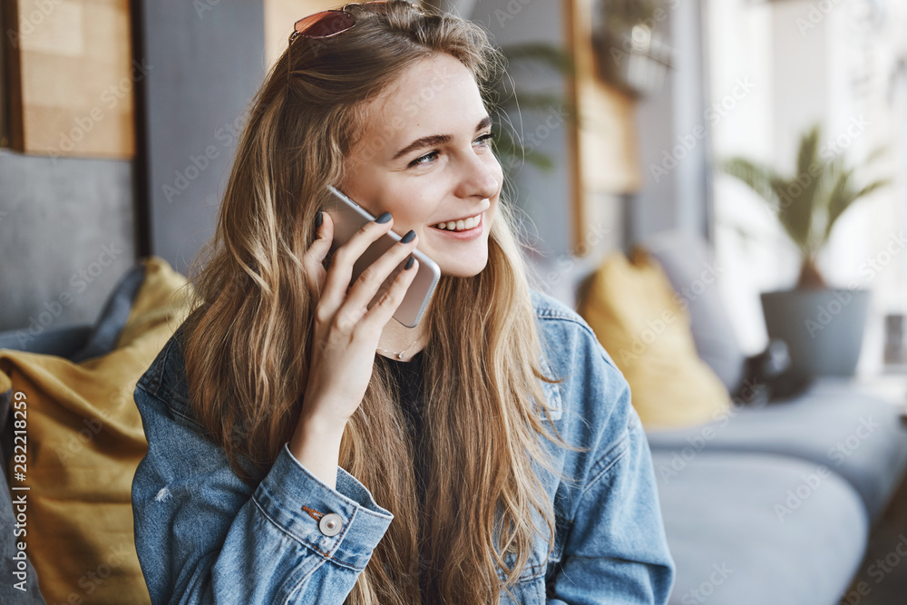 Good-looking happy fair-haired girlfriend in stylish denim jacket, looking aside at street and smiling while talking on smartphone, calling friend while waiting for her in cafe, resting and chilling Good-looking happy fair-haired girlfriend in stylish denim jacket, looking aside at street and smiling while talking on smartphone, calling friend while waiting for her in cafe, resting and chilling
