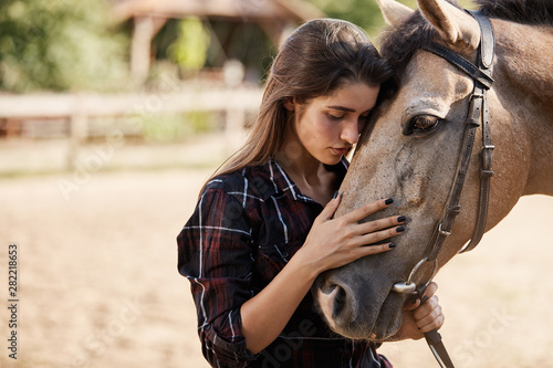 Close-up tender attractive caucasian brunette woman hugging brown horse, petting its nose, close eyes lean and touching animal foretop, enjoy spend time on farm in meadow, owner lead it to stable photo