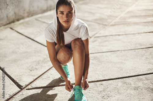 Motivation, urban fitness and running concept. Determined alluring young fit female workout outdoors, kneel to tie shoelaces on sneakers, look camera self-assured, prepare morning jogging