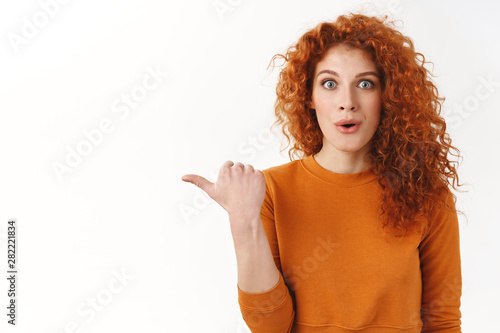 Astonished impressed redhead curly woman pointing thumb left, showing blank white copy space, fold lips amused stare camera charmed, discuss amazing product, standing white background
