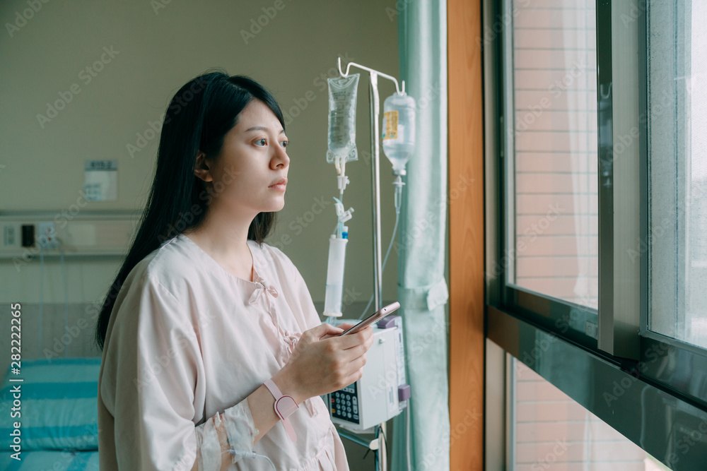 young woman at hospital room wearing patient pajamas standing by window holding mobile phone. unhappy girl talking about health condition online worried and concerned. sickness lady look out window
