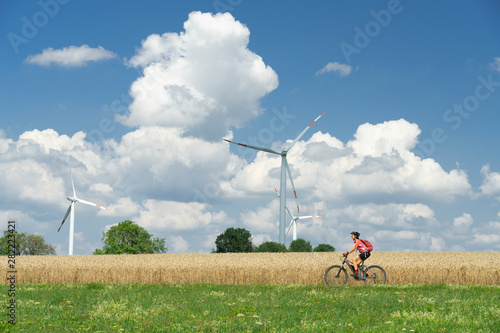 nice, active woman, riding her electric mountain bike between wheat fields and wind wheewls of a wind farm on the Schwaebische Alb near the city of Aalen, BadenWuerttemberg, Germany photo