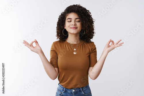 Om girl feels peace patience. Attractive carefree relaxed happy young woman curly shirt hairstyle close eyes smiling delighted meditating hands sideways nirvana lotus pose, breathing practice yoga photo