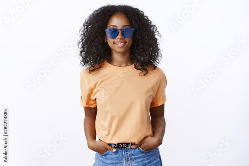 Attractive cool stylish african-american girl wearing sunglasses orange t-shirt holding hands pockets confident, smiling carefree enjoying sunbeams summer walk friends, self-assured look fashionable