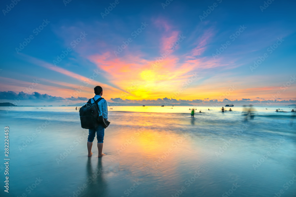 Fototapeta premium Silhouette of man backpacker on the beach looking at magical dramatic sunrise. The man standing on the sandy beach