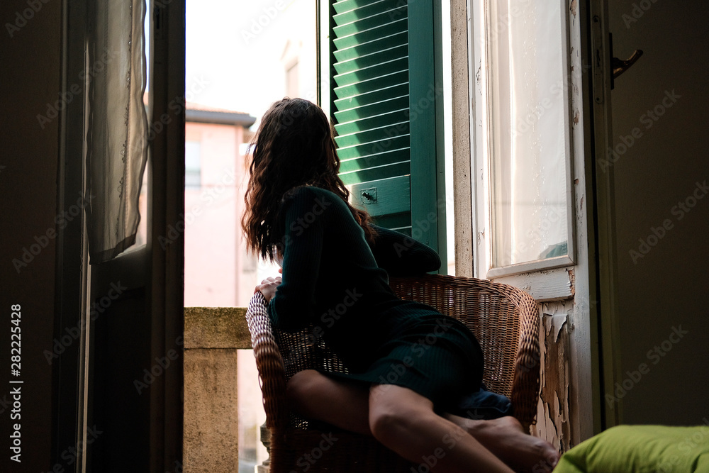 Woman in tight knit dress sitting in wicker chair on a balcony with green wooden shutters in traditional italian hotel. Female solo travel concept