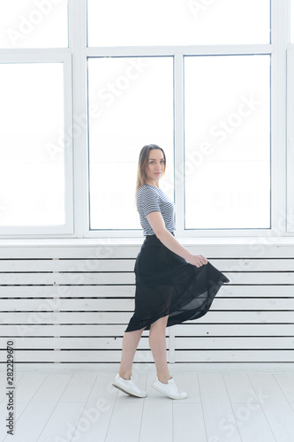 Youth, style and people concept - young woman on black skirt and sneakers standing near the window