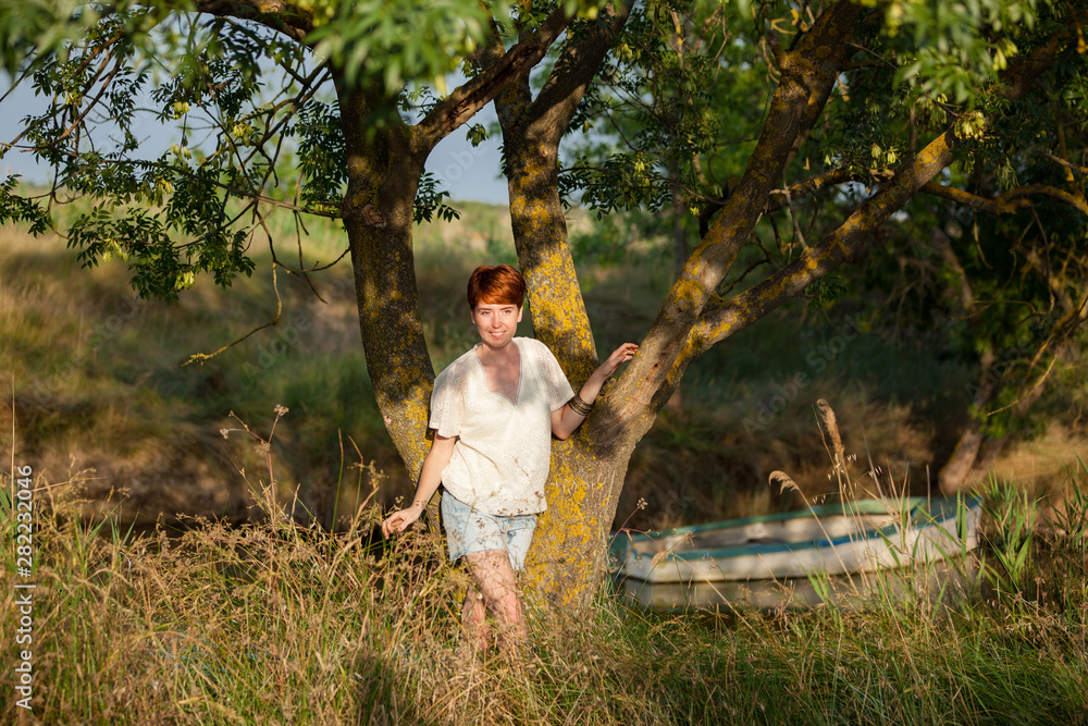WELL-BEING, WOMAN AGAINST A TREE IN THE GRASS IN THE COUNTRYSIDE.