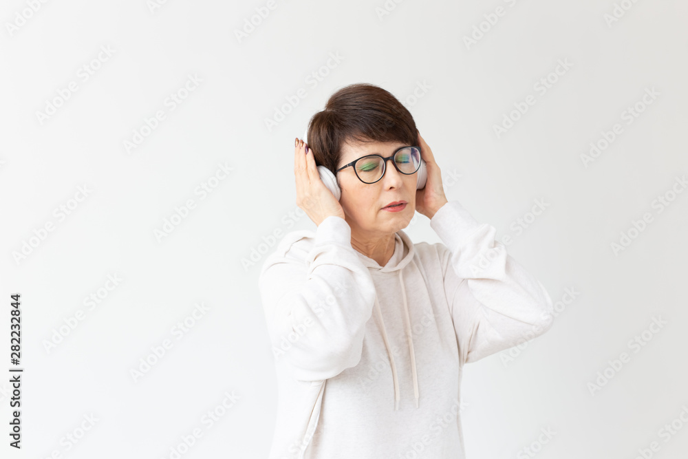 Cute positive middle-aged woman in sweater and glasses is listening to music with wire headphones standing on a white background. Concept of hobbies and subscriptions to favorite radio station.