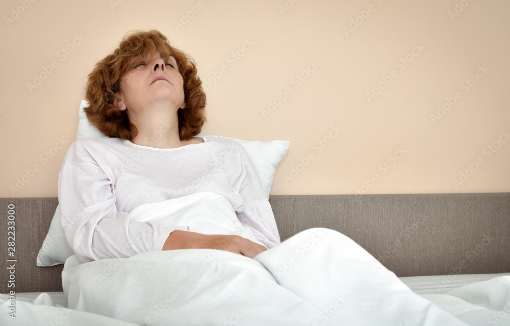 Tired and exhausted mature woman with closed eyes sitting in bed leaning on the pillow Tired and exhausted mature woman with closed eyes sitting in bed leaning on the pillow