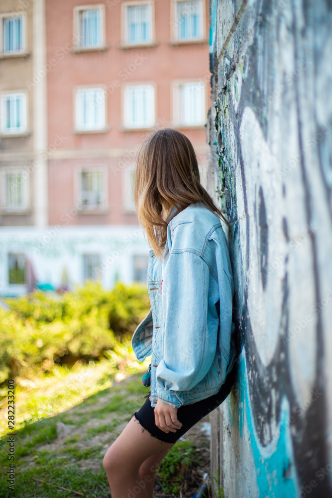 Pretty young female in casual outfit and leaning on weathered graffiti wall while standing on city street on sunny day