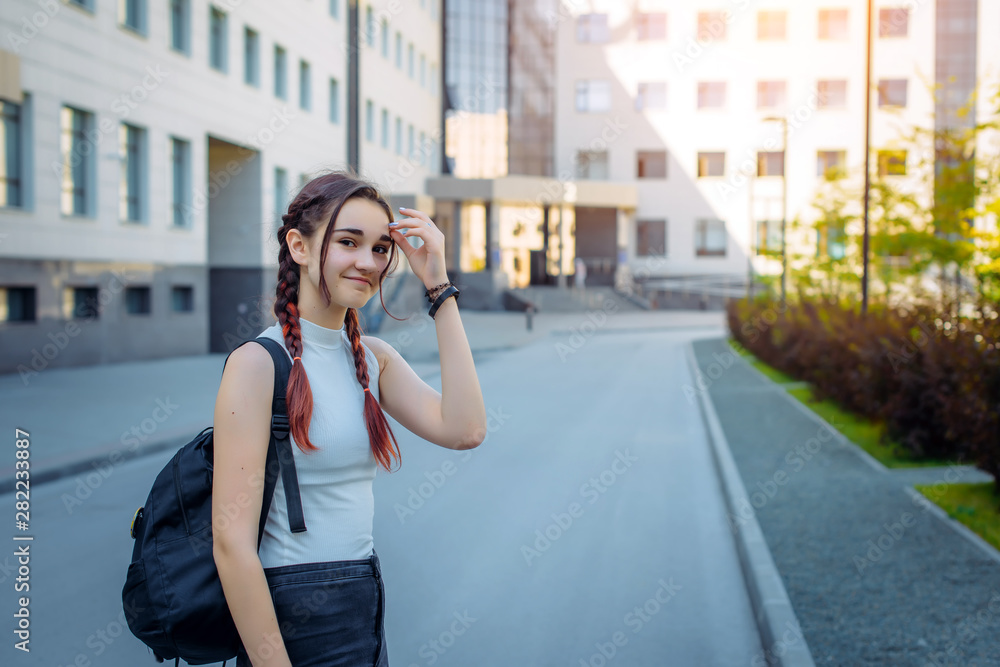 Young beautiful student with backpack on her shoulder goes to school, close-up. Schoolgirl with two braids standing in front of college, blurred background. September 1, the beginning of school year.