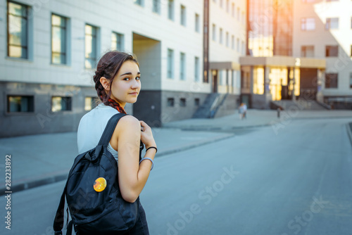 Rear view beautiful brunette with a black backpack behind going to College. A young pretty student looks back at the camera. A beautiful girl going to school. Concept of the Day of knowledge.