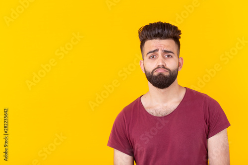 Sad young handsome man hipster with a beard in a burgundy t-shirt posing on a yellow background. Concept of frustration.