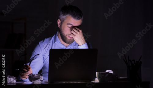 Tired man is working on laptop in darkness