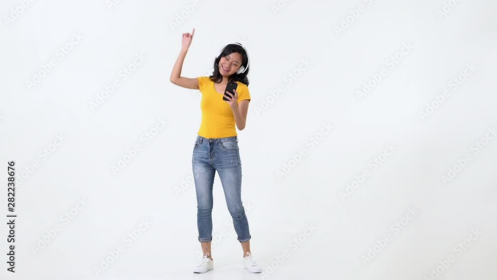 Smiling beautiful young asian woman holding mobile phone in hand enjoying the music on headphones against white background Smiling beautiful young asian woman holding mobile phone in hand enjoying the music on headphones against white background
