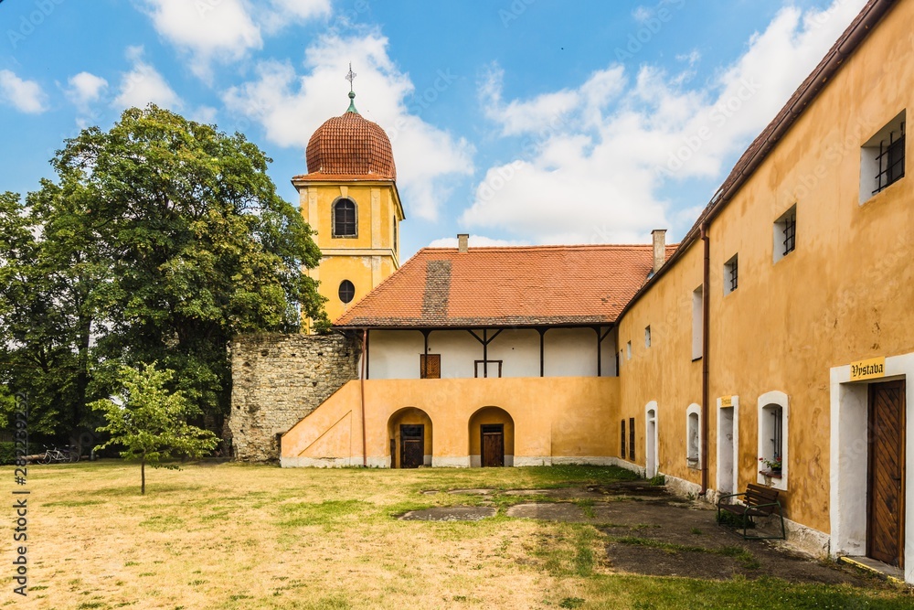 Panensky Tynec, Czech Republic - July 15 2019: Yellow bell tower and former monastery of Poor Clares, now the municipal authority. Sunny day with blue sky and clouds. View from garden with grass.