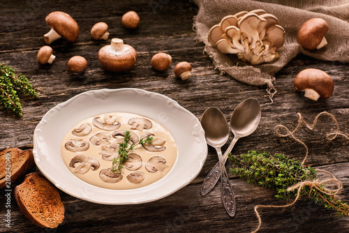 Fototapeta Plate with mushroom cream soup, pieces of toasted bread, melchior spoons, bundle of fresh thyme, brown royal champignons and oyster mushrooms on wooden table