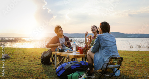 A group of Asian friends tourist drinking and playing guitar together with happiness in Summer while having camping near lake at sunset
