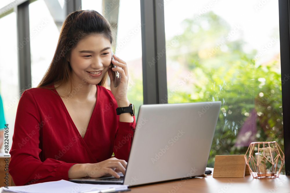 Attractive Asian woman wearing red dress working with laptop smile and talking with customer in smartphone to get idea and requirement for success job outside office in coffee shop,Business Concept Attractive Asian woman wearing red dress working with laptop smile and talking with customer in smartphone to get idea and requirement for success job outside office in coffee shop,Business Concept
