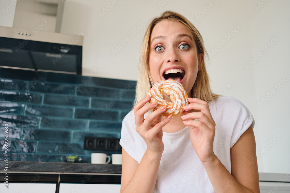 Young cheerful blond woman in white T-shirt holding sweet bun for breakfast in hands amazedly looking in camera at modern cozy home Young cheerful blond woman in white T-shirt holding sweet bun for breakfast in hands amazedly looking in camera at modern cozy home