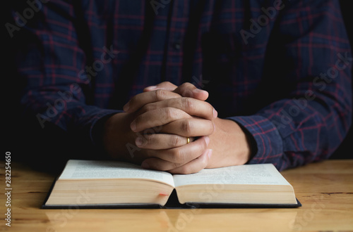 Hands of a young man folded praying over a Bible on wooden table