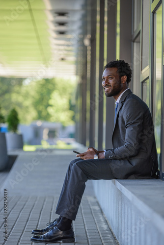 cheerful african american american businessman using smartphone and looking away while sitting on parapet