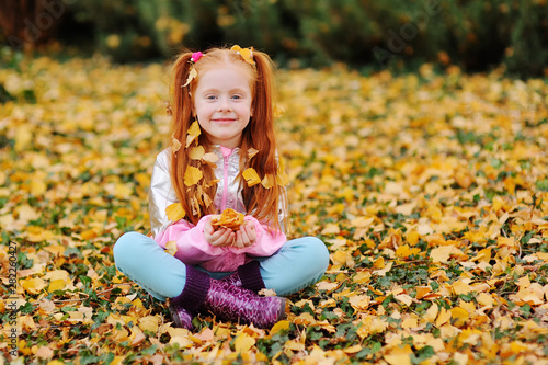 little cute red-haired girl child with yellow fallen leaves on her hair smiling against the autumn leaves in the Park.space for text, copy space,