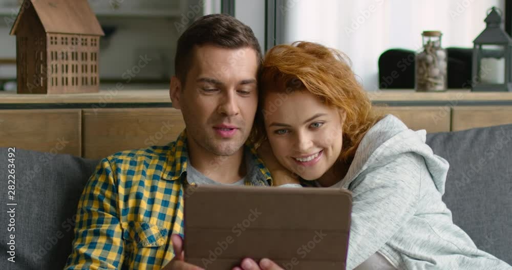 Happy young couple has video chat with their friends, family members, holding tablet computer, smiling, looking at webcamera, sitting on grey sofa at home. 4K, shot on RED camera. Happy young couple has video chat with their friends, family members, holding tablet computer, smiling, looking at webcamera, sitting on grey sofa at home. 4K, shot on RED camera.