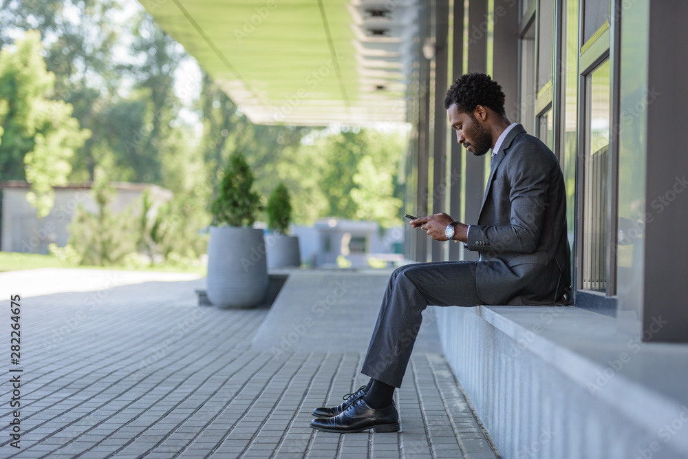 thoughtful african american businessman using smartphone while sitting on parapet thoughtful african american businessman using smartphone while sitting on parapet
