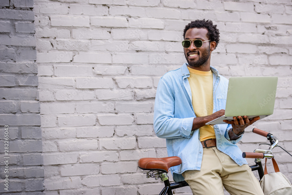 Fototapeta premium smiling african american man looking away while standing by brick wall near bicycle and using laptop
