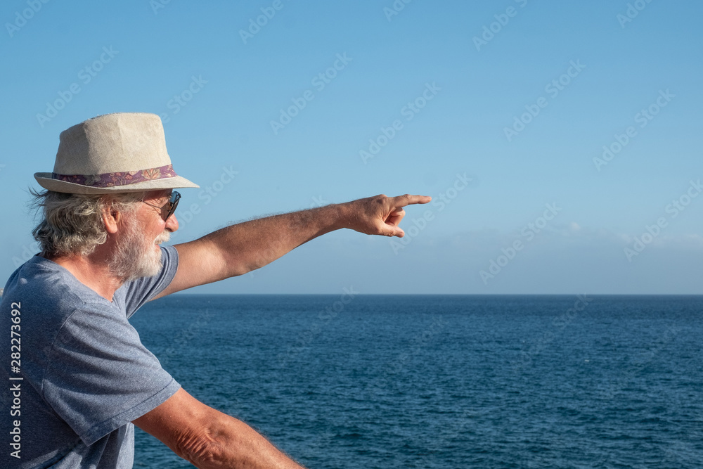 Active senior man smiling in front of the sea. Blue sky and sunny day. With the arm indicates a distant point. Horizon over water
