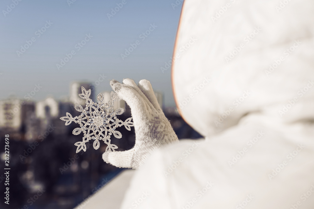 Woman in a jacket and knitted gloves holding huge sparkling snowflake on the town background. Winter and Christmas concept. Woman in a jacket and knitted gloves holding huge sparkling snowflake on the town background. Winter and Christmas concept.