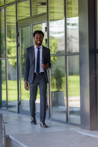 smiling african american businessman holding smartphone walking near office building