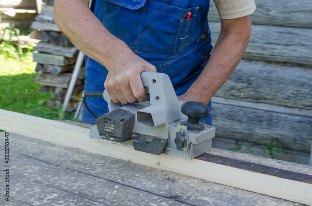 The hand of a man working with a planer and planer boards. Carpenter working with electric planer on wooden plank outdoor. Processing of material. Work tool. Soft focus. The hand of a man working with a planer and planer boards. Carpenter working with electric planer on wooden plank outdoor. Processing of material. Work tool. Soft focus.